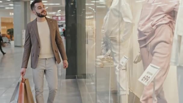 Střední záběr pohledný vousatý muž v chytrém oblečení nesoucí spoustu nákupních tašek kráčející podél nákupního centra při pohledu na figuríny v okně displeje - Záběry, video