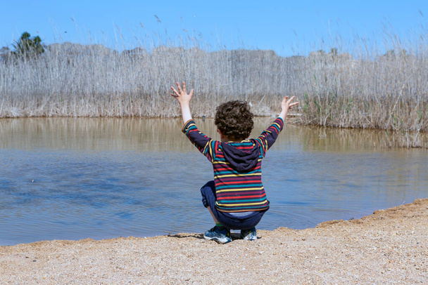 A kisfiú a tó szélén guggolt göndör hajjal és színes csíkos pólóval, feltartott karokkal játszott.. - Fotó, kép