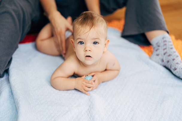 Winziges Baby in Windeln liegt mit dem Bauch auf einer blauen Decke und hält ein Spielzeug in den Händen. Mütterhände halten das Baby am Rücken - Foto, Bild