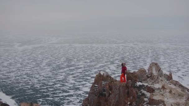 Mies punaisessa takissa seisoo vuorella jääpeitteisen Baikal-järven takana. Vapaus ja vahvuus käsite - Materiaali, video