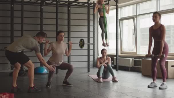 Široký záběr žen, které cvičí v tělocvičně, dřepí s činkami a činkami, sedají si na podložku na jógu na podlaze, zvedají se s kapelami. Mužský trenér pozorování a pomáhá udržovat techniku - Záběry, video