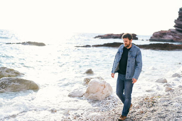 Pensive man in jeans and a denim jacket walks along a pebble beach near the water looking under his feet against a background of rocks - Fotó, kép