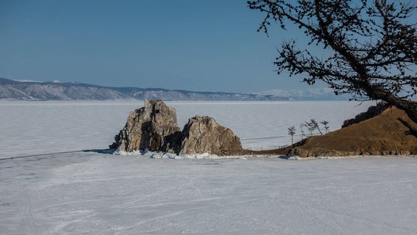 Kaksipäinen kivi, jolla ei ole kasvillisuutta, nousee jäätyneen järven yläpuolelle, ja jäällä näkyy pieniä siluetteja ihmisistä. Paljaat oksat puun taustalla sininen taivas. Talvipäivä. Baikal - Valokuva, kuva