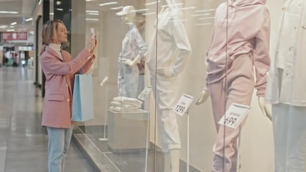 Середній знімок молодої красивої жінки в розумному повсякденному одязі, що стоїть біля вікна, демонструє модний магазин одягу, фотографуючи манекен на смартфоні
 - Кадри, відео