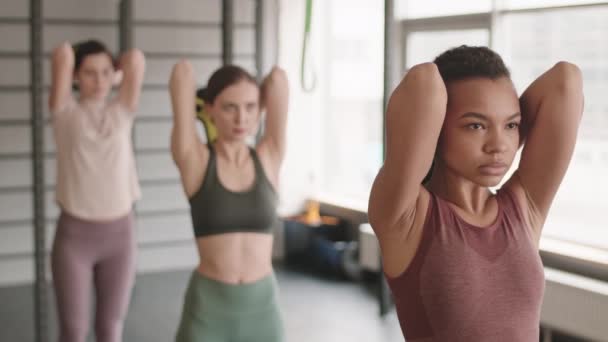 Pas-up młodej mieszane rasy kobieta robi triceps rozszerzeń ze sztangą wagi wraz z rozmytych sportowców stojących w jednym rzędzie za nią w siłowni - Materiał filmowy, wideo