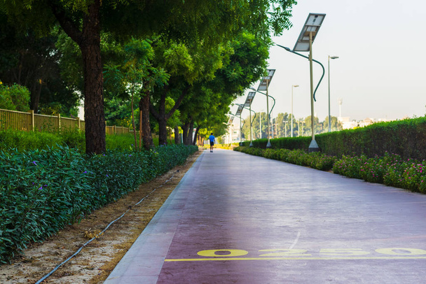 Homme d'âge moyen marchant sur la piste de jogging dans le parc Nad Al Hamar, tôt le matin. Le poteau de lampe alimenté par des panneaux solaires peut être vu dans l'image. - Photo, image