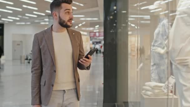 Naklápěcí střední záběr pohledný vousatý muž v chytré ležérní oblečení focení stylové oblečení na figuríny v okně displej módní oblečení obchod ve velkém nákupním centru - Záběry, video