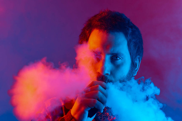 portrait of bearded man vaping with colorful neon lights. person quitting smoking with substitution therapy. boy looking at camera - Foto, Imagen