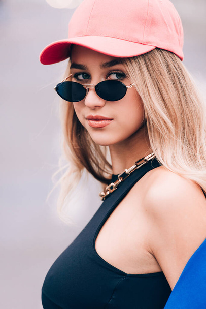 Молода сексуальна блондинка хіпстер позує на вулиці. Носіння синього стильного піджака, джинсів і бейсбольного капелюха і сонцезахисних окулярів
. - Фото, зображення