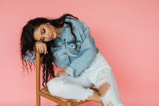 Emotional and psychological problems. Sad and disappointed girl sits on a chair in denim jacket on pink background. - Photo, image
