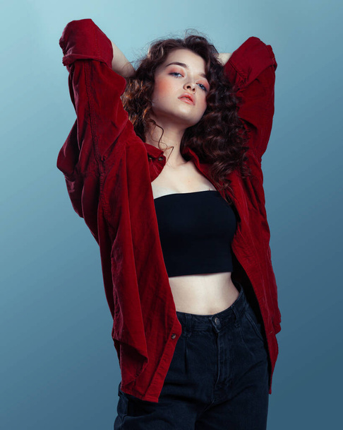 Attractive brunette with long curly hair. Female fashion model with wavy hairstyle in a red shirt posing on a blue background - Fotografie, Obrázek
