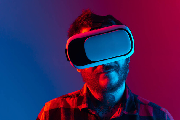Χαμογελώντας άνθρωπος με συσκευή εικονικής πραγματικότητας στο κεφάλι του. Studio πορτρέτο με φώτα νέον. άτομο που χρησιμοποιεί γυαλιά VR. - Φωτογραφία, εικόνα