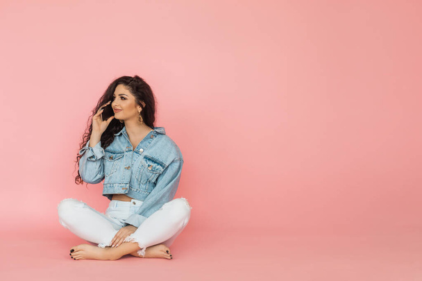 Jovem bela estudante em roupas jeans em fundo rosa no estúdio senta-se de pernas cruzadas no chão. Usando o telefone celular para se comunicar. - Foto, Imagem