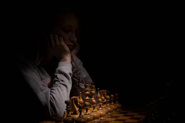 the girl looks thoughtfully at the chessboard against a dark background. old chess. business concept - Foto, afbeelding