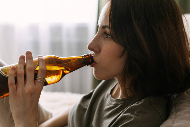 Egy fiatal nő sört iszik, miközben a kanapén ül. Női alkoholizmus - Fotó, kép