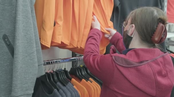 Mujer caucásica en máscara contra el virus elige una camisa naranja para comprar en una tienda - Metraje, vídeo
