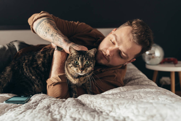 Mann streichelt seine gestreifte Katze, während er Zeit mit ihr im Schlafzimmer verbringt - Foto, Bild
