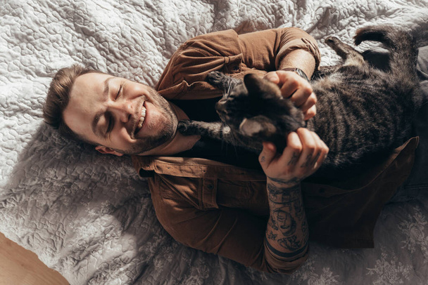 Man laughing and stroking his striped cat while spending time with him at bedroom - Фото, изображение