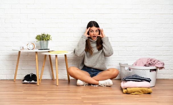 Junge Frau sitzt zu Hause mit Wäschekorb mit überraschtem Gesichtsausdruck auf dem Boden - Foto, Bild