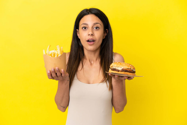 Junge kaukasische Frau hält Burger und Chips isoliert auf gelbem Hintergrund - Foto, Bild