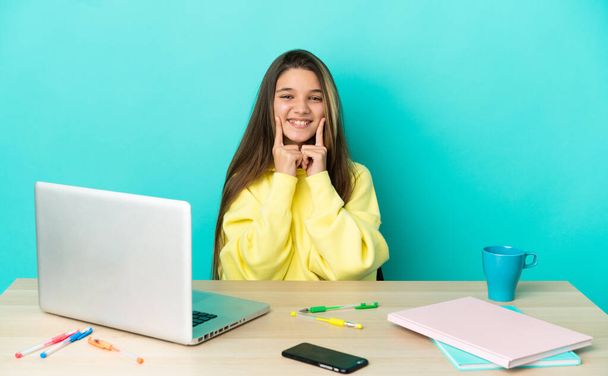 幸せと快適な表情で笑顔を隔離された青い背景の上にノートパソコンを持つテーブルの少女 - 写真・画像