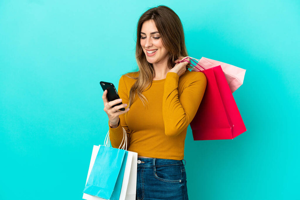 Mulher caucasiana isolada no fundo azul segurando sacos de compras e escrevendo uma mensagem com seu telefone celular para um amigo - Foto, Imagem