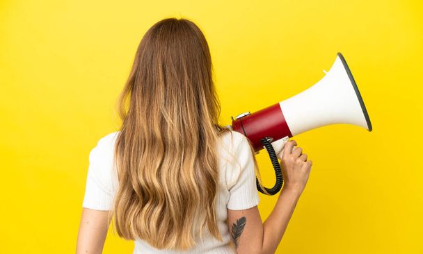 Caucasian woman isolated on yellow background holding a megaphone and in back position - Photo, Image