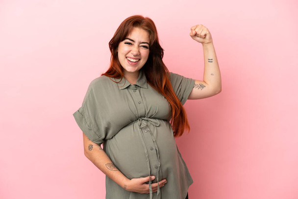 Молодая рыжая белая женщина изолирована на розовом фоне беременной и делает сильный жест - Фото, изображение