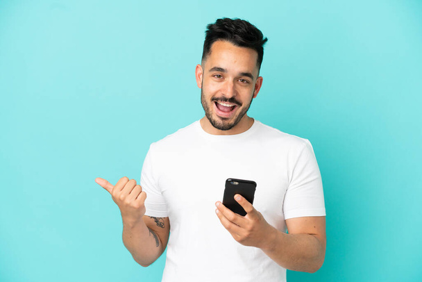 Nuori valkoihoinen mies eristetty sinisellä pohjalla matkapuhelimella ja osoittaa sivusuunnassa - Valokuva, kuva