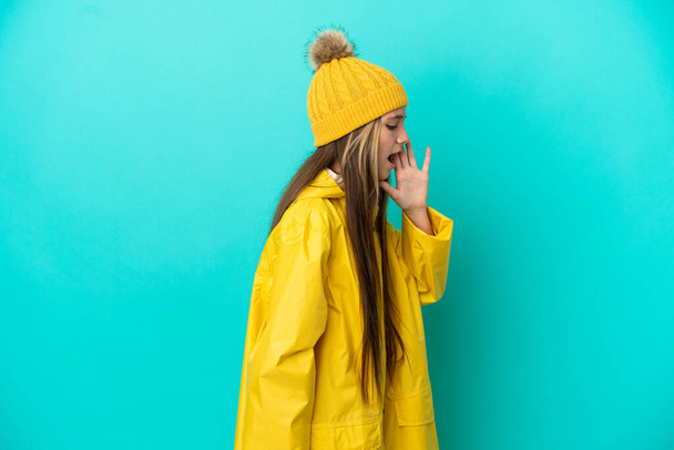 Pikkutyttö yllään sateenkestävä takki yli eristetty sininen tausta huutaa suu auki sivulle - Valokuva, kuva