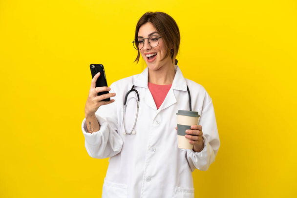 Médica mulher isolada no fundo amarelo segurando café para levar e um celular - Foto, Imagem
