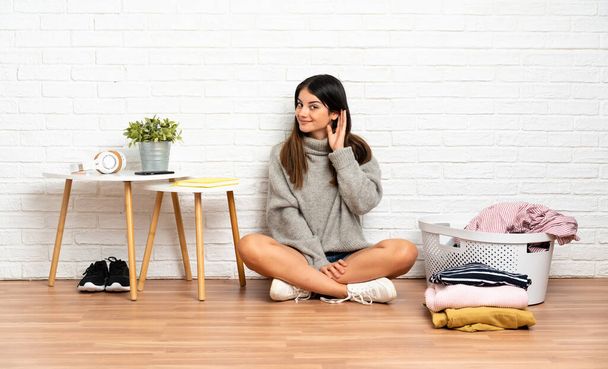 Jeune femme assise sur le sol à l'intérieur avec panier à vêtements écoutant quelque chose en mettant la main sur l'oreille - Photo, image