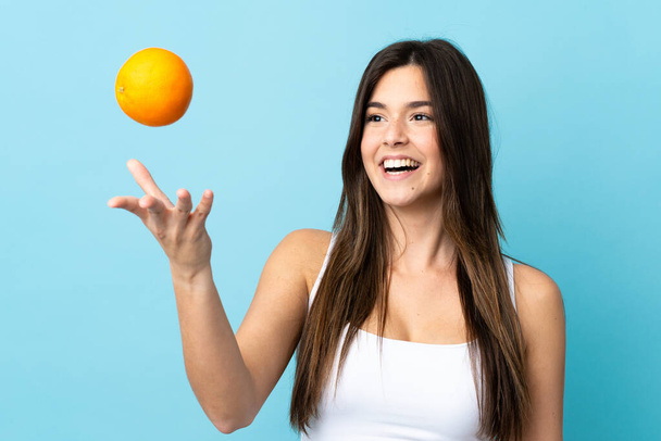 Teenager Brazilian girl over isolated blue background holding an orange - Fotoğraf, Görsel