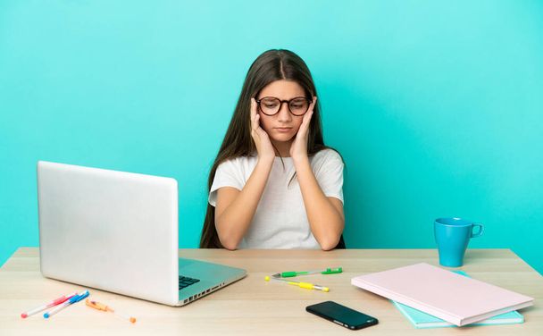 Menina em uma mesa com um laptop sobre fundo azul isolado com dor de cabeça - Foto, Imagem