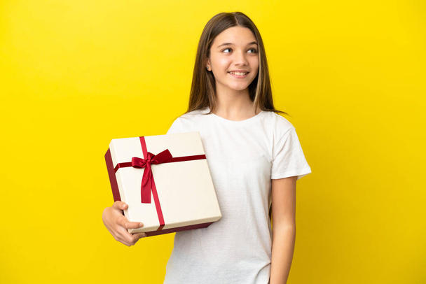 Kislány kezében egy ajándék felett elszigetelt sárga háttér gondolkodás egy ötlet, miközben felnéz - Fotó, kép