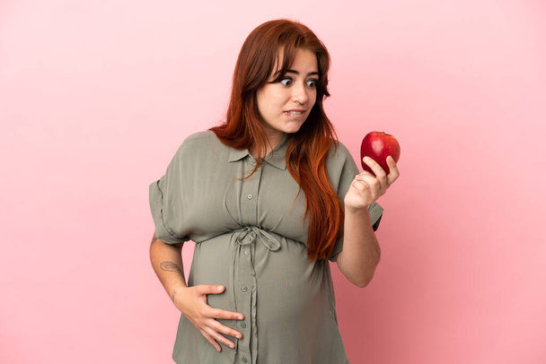 Νεαρή κοκκινομάλλα καυκάσια γυναίκα απομονωμένη σε ροζ φόντο έγκυος και κρατώντας ένα μήλο - Φωτογραφία, εικόνα
