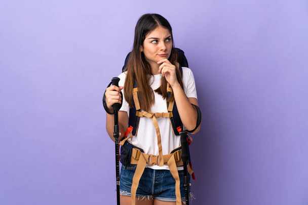 Young woman with backpack and trekking poles isolated on purple background having doubts while looking up - Fotografie, Obrázek