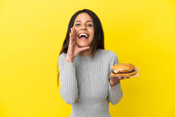 Young caucasian woman holding a burger isolated on yellow background shouting with mouth wide open - Fotoğraf, Görsel