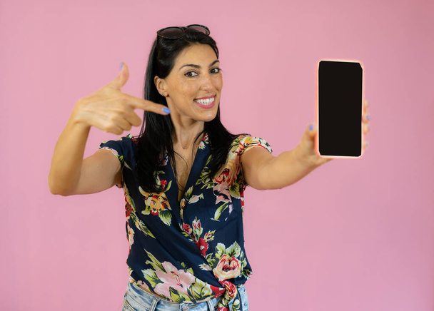 Smiling woman is pointing on smartphone standing over pink background. - Фото, изображение