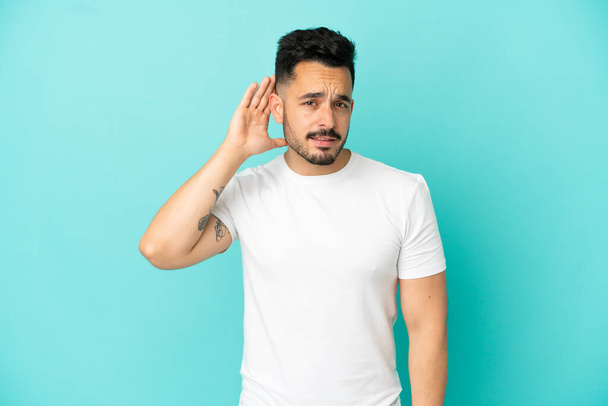 Young caucasian man isolated on blue background listening to something by putting hand on the ear - Foto, Bild