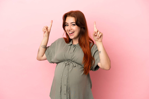 Junge rothaarige kaukasische Frau isoliert auf rosa Hintergrund schwanger und zeigt nach oben - Foto, Bild