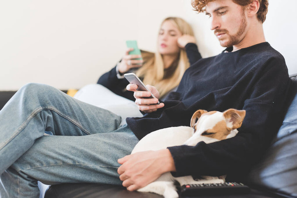 若いです男座ってソファ屋内でホーム使用スマートフォンとともにジャックラッセル犬スリープオン彼の腕 - 写真・画像
