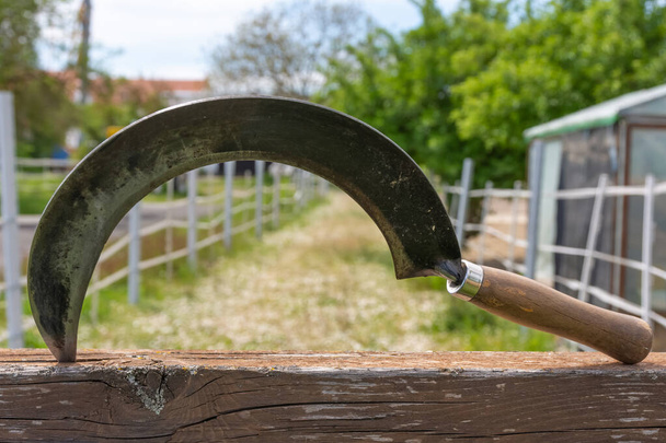 A closeup shot of a sickle stuck on a plank of wood on a background of a rural landscape - Foto, imagen