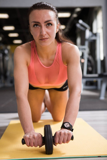 Κάθετη λήψη μιας αθλήτριας που δυναμώνει τους μυς του πυρήνα της, χρησιμοποιώντας τροχό ab στο γυμναστήριο - Φωτογραφία, εικόνα