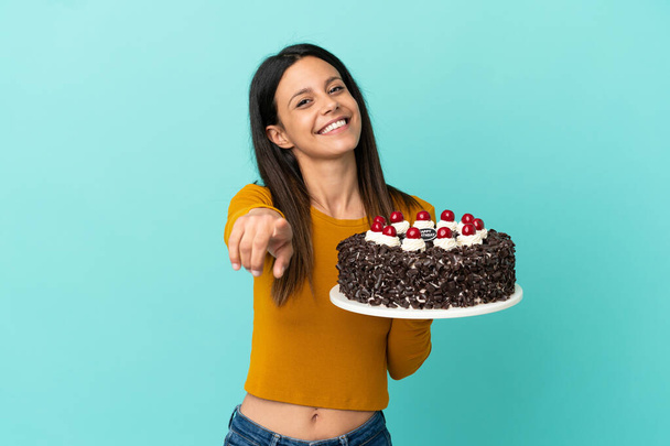 Mladá běloška drží narozeninový dort izolované na modrém pozadí ukazuje dopředu s šťastným výrazem - Fotografie, Obrázek
