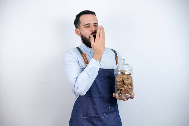 Fiatal férfi szakállal elszigetelt fehér háttér kezében egy üveg csokoládé chips cookie unatkozik ásítás fáradt takaró száj kézzel. Nyugtalan és álmos. - Fotó, kép