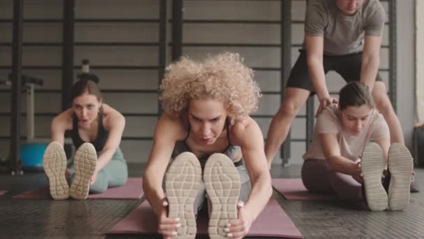 Full shot of three slim Caucasian women sitting on yoga mats in gym, doing seated forward bend, male fitness coach helping them - Imágenes, Vídeo