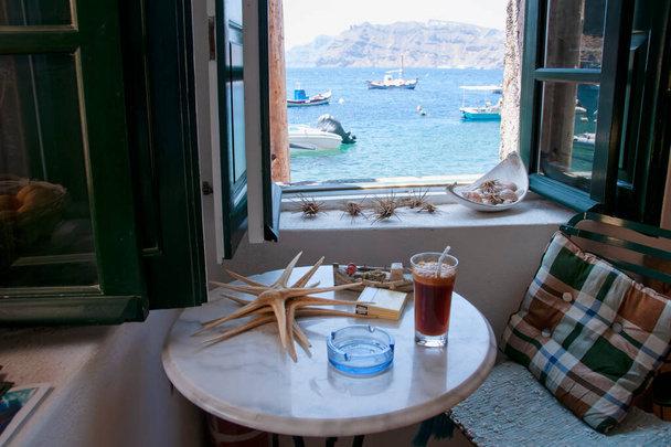 A view from the sea window in Amoudi, the cozy fisherman's house with starfish and a drink on the table. In the background the caldera of the volcano - Photo, Image