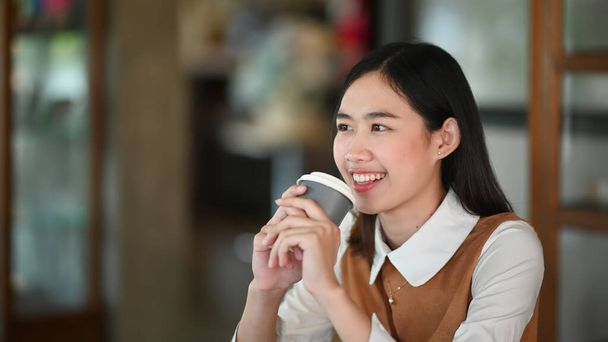 Веселая молодая женщина улыбается и держит чашку кофе, сидя в кафе. - Фото, изображение