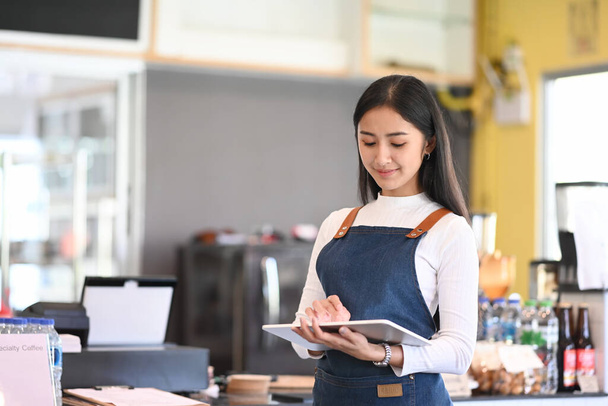 Young woman coffee shop owner wearing apron holding digital tablet ready to receive orders. - Photo, Image